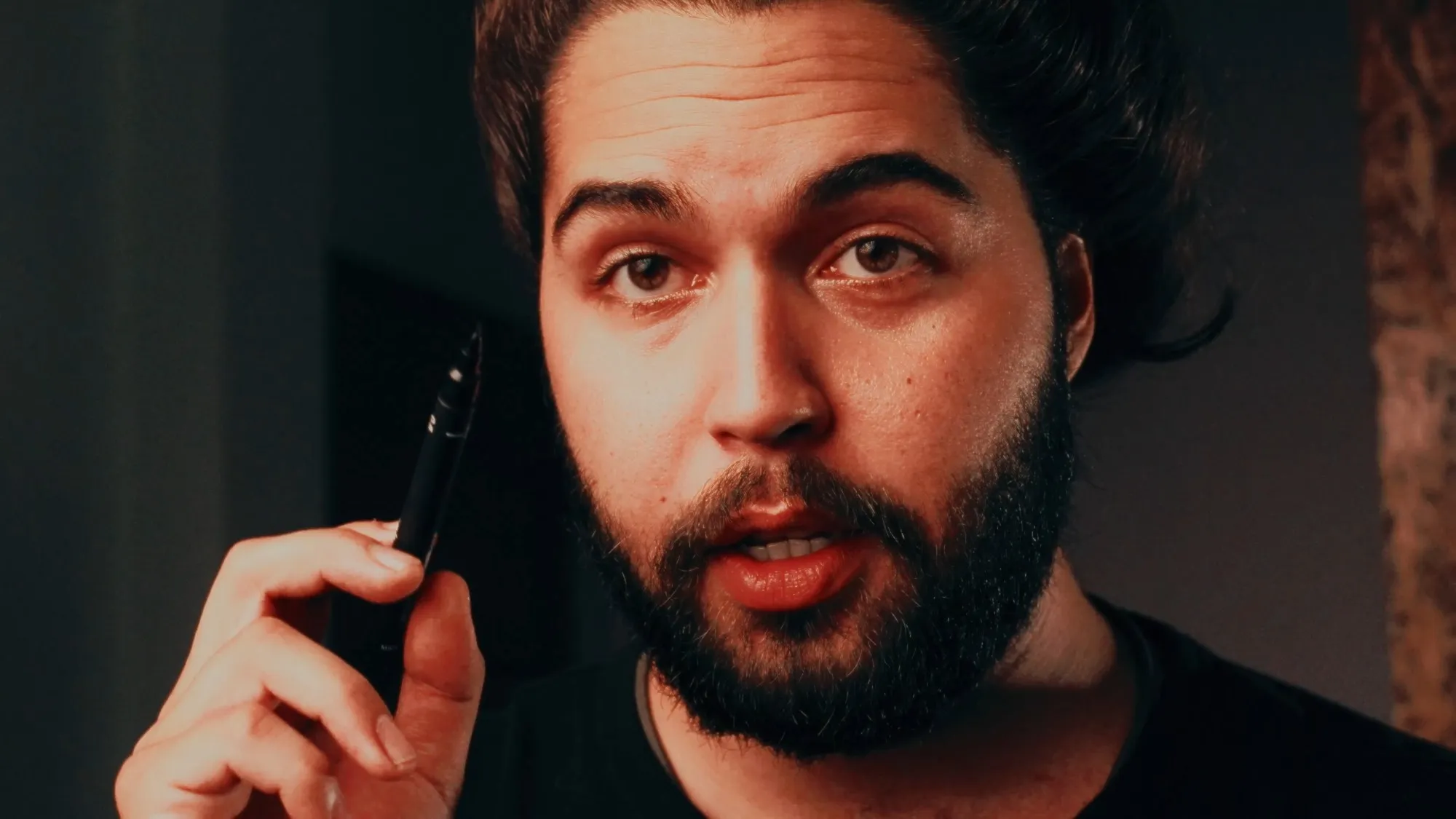 Close-up of a bearded man mid-sentence, holding a pen up near his face